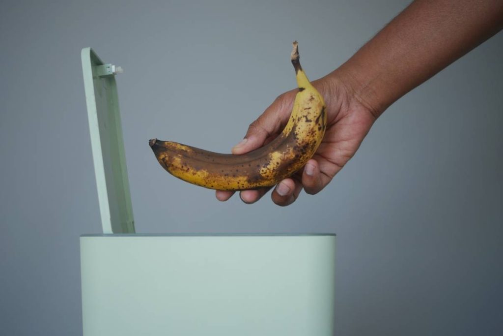 A person throws away a brown banana into a green trash can.